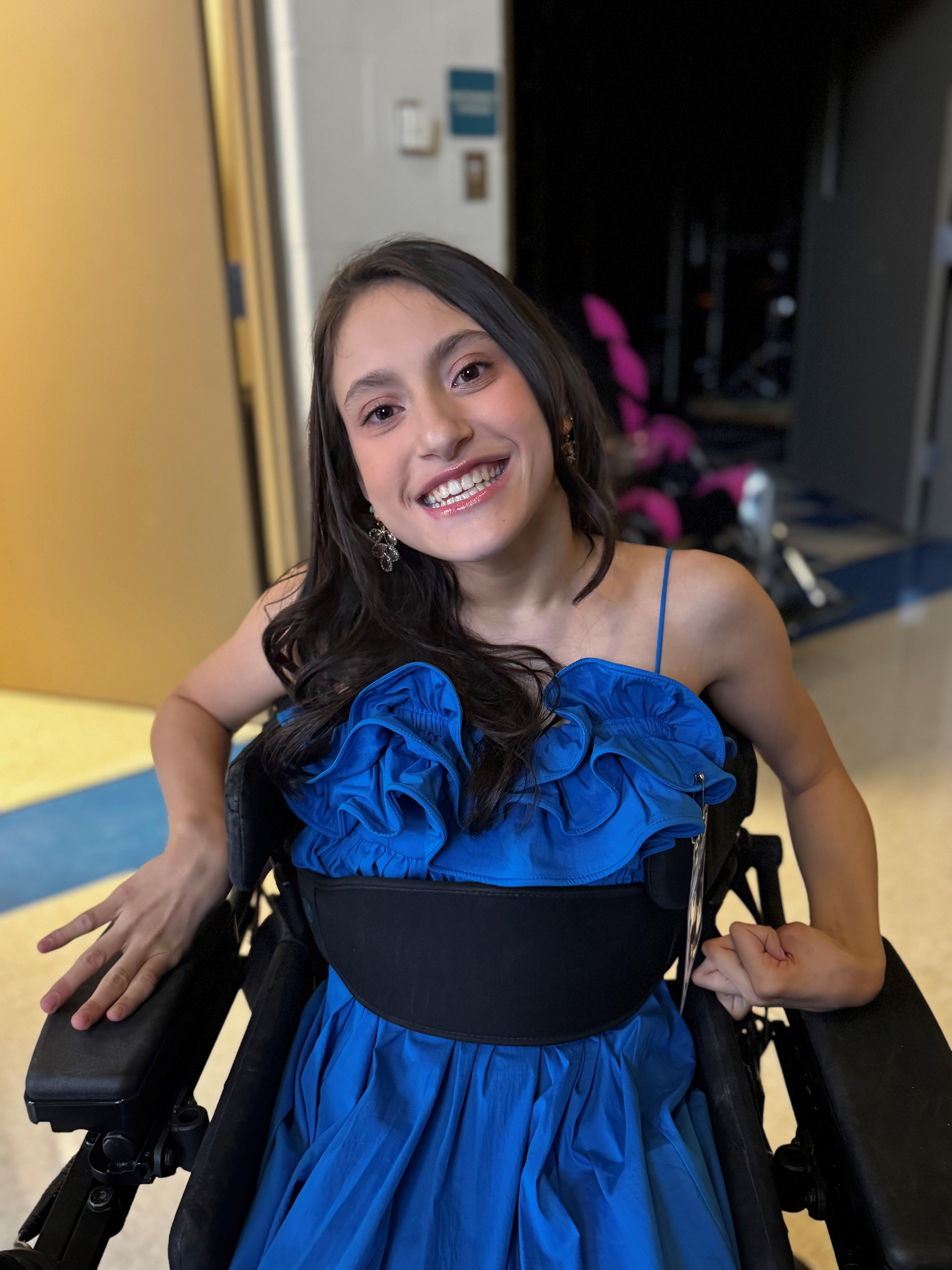 A beautiful young woman with long, gorgeous hair and a radiant smile sits in a wheelchair, wearing a pretty blue dress.