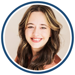 A headshot of a woman with long, wavy brown hair with blonde highlights, smiling at the camera. She is wearing a soft pink top. The image is framed within a circular border with a blue outline. The background is a light, neutral color.