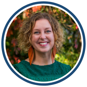 A headshot of a woman with curly, shoulder-length blonde hair, smiling at the camera. She is wearing a green textured blouse and large, orange feather-shaped earrings. The image is framed within a circular border with a blue outline. The background features blurred autumn foliage with red and green leaves.