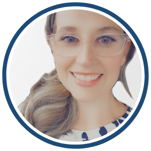  circular headshot of a woman with light brown hair styled in a side ponytail, wearing clear-framed glasses and a black-and-white patterned blouse. She has a bright smile and is set against a clean, white background. The image is framed with a dark blue border.