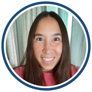 A circular headshot of a smiling woman with long, straight, dark brown hair, wearing a pink top. The background features light-colored curtains. A dark blue border surrounds the image.