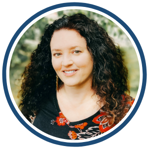 A circular headshot of a woman with long, curly dark hair, smiling with a natural outdoor background. She is wearing a black top with a colorful red and orange floral pattern. The image is framed with a dark blue border.