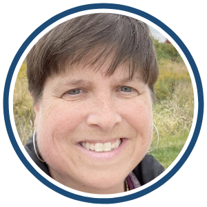 A circular headshot of a woman with short brown hair and blue eyes, smiling outdoors with a natural landscape in the background. She is wearing a black jacket and silver hoop earrings. The image is framed with a dark blue border.